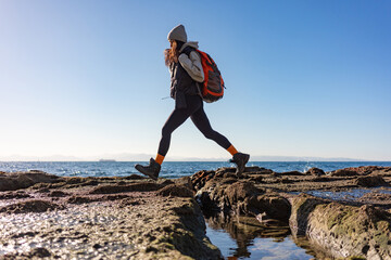 Active female hiker with orange backpack jumping over rocky coastal terrain at low tide, dynamic seaside adventure scene with motion, balance, freedom, ocean horizon, and copy space sky