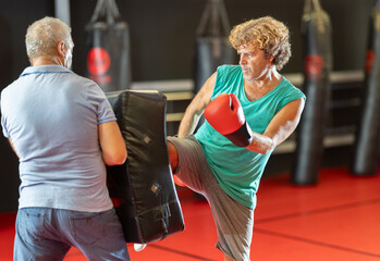 Focused middle-aged man in red boxing gloves landing fast waist-level kick against padded shield, practicing distance control and power with older coach in self-defense session