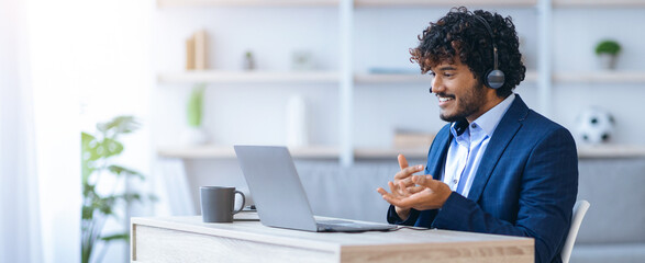 A man sits at a desk wearing a suit and headset. He is engaged in a conversation on his laptop. Bright light fills the room, showing a modern office with plants and a soccer ball.