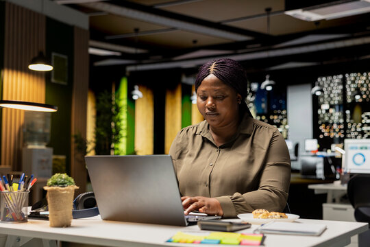 Young businesswoman spends night time at the office eating unhealthy food, dealing with a stress eating disorder due to hard work after hours. Emotional compulsive overeating at the firm.