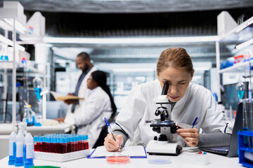Fototapeta premium Quality control analyst examining bacterial cultures on slide using lab microscope. Woman ensuring consumer safety by checking for microscopic pathogens with optical device