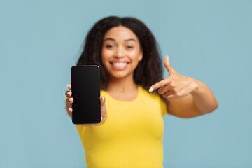 Fototapeta premium Mockup image of black lady pointing at smartphone with blank screen, demonstrating free place for design or advertisement, recommending app or website, blue studio background