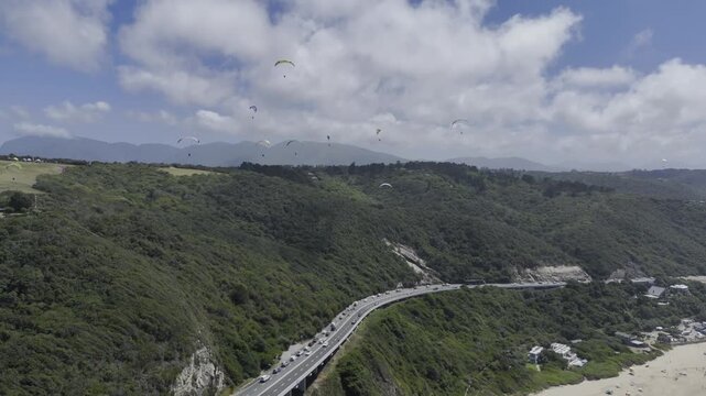 Drone flies east along coastal highway as paragliders fill the sky above on a sunny day on the Garden Route in Wilderness, South Africa