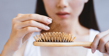 A woman sits indoors, holding a hairbrush in one hand and strands of hair in the other. She looks focused as she examines the hair caught in the brush.