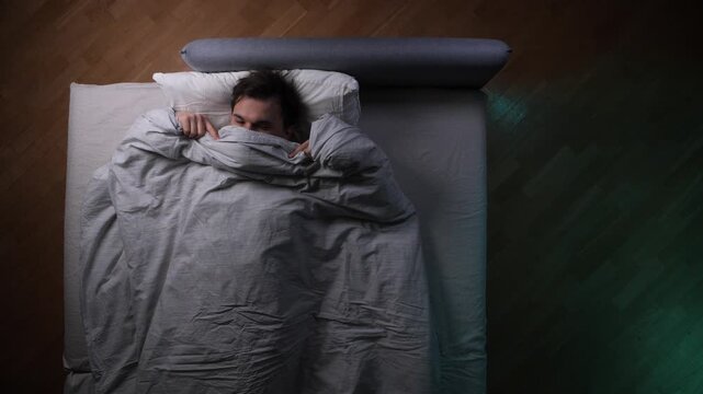 Young man awake in bed at night, pulling the blanket over his face, suffering from insomnia, anxiety, and sleep problems, feeling restless and unable to fall asleep
