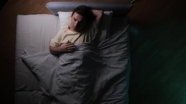 Young man restlessly lying awake in bed, struggling with insomnia and sleeplessness at night, feeling anxious and stressed about not being able to fall asleep and experiencing mental health issues