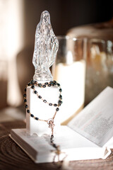 A clear statue of a religious figure stands on a table next to an open Bible. A rosary made of black beads is resting on the Bible, creating a simple but meaningful scene.