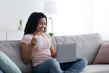New normal, covid-19 outbreak, work at home, remote lesson, study and education, video call. Cheerful millennial african american lady typing on laptop, hold cup of drink, in living room interior