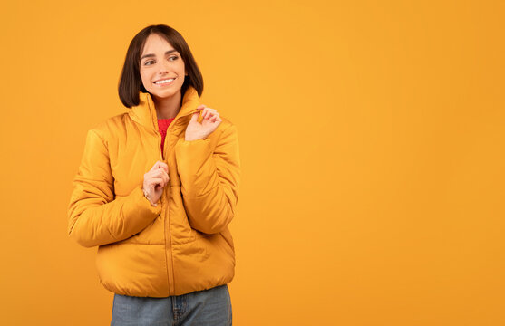 Autumn clothes concept. Excited lady wearing and zipping up jacket, smiling and looking at empty space, standing over orange studio background, free space