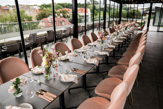 Long dining table arranged with floral accents, soft textiles, and modern tableware inside a bright glass space prepared for a private gathering or social event.