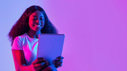 A joyful young black woman is sitting in a neon-lit space, using a tablet to study or work. She appears engaged and excited during her online meeting, showcasing modern remote work culture.