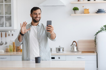 Cheerful handsome adult european male waving hand to smartphone webcam, making video calls from phone, communicating remotely on minimalist kitchen interior. Meeting remotely, social distance and app