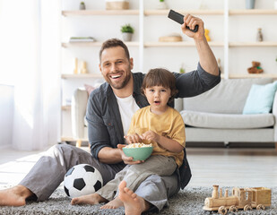 Family of soccer fans. Little boy watching football game on tv, supporting favorite team, sitting on floor together with ball and popcorn, free space © Prostock-studio