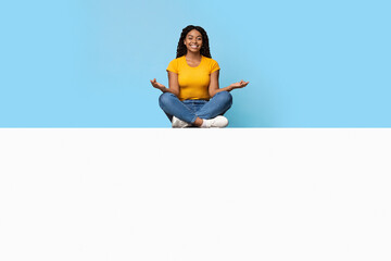 Positive pretty young african american lady in casual sitting in lotus pose and smiling, meditating on white horizontal empty board for text or advertisement, blue studio background, copy space
