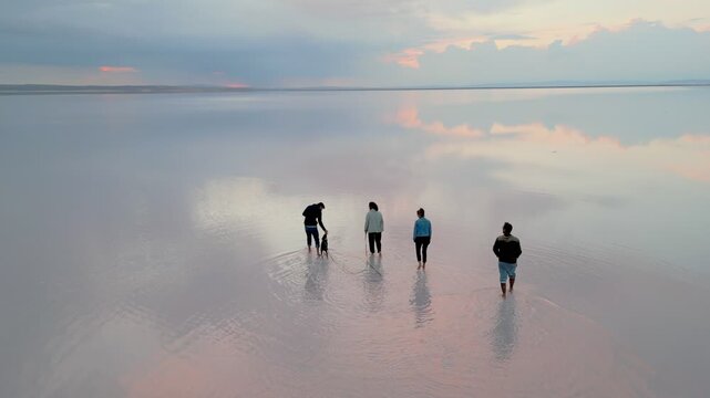 Friends walking barefoot with a dog on the salt flat reflecting the cloudy sky at sunset drone aerial view in Turkey