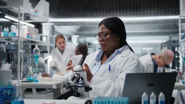 Female molecular biologist studying genetic tissue sample on glass slide under magnification. African american woman searching for mutation patterns using advanced optical equipment, camera A