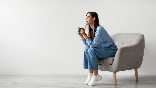 Time for coffee. Charming millennial woman sitting in armchair, enjoying cup of hot drink against white studio wall, banner with free space. Young lady relaxing with mug of warm beverage