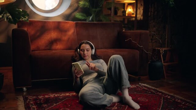 Young woman lying on the floor at home, scrolling smartphone and yawning. Cozy evening atmosphere, fatigue after long day, social media browsing, casual lifestyle and relaxation concept