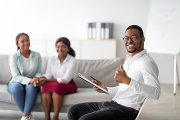 Happy black male psychologist showing thumb up gesture, young lesbian couple holding hands and...