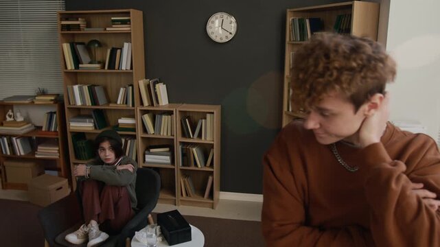 Upset teenage boy standing by window with arms crossed unwilling to talk to girlfriend holding grudge against partner during couple therapy in counselling office