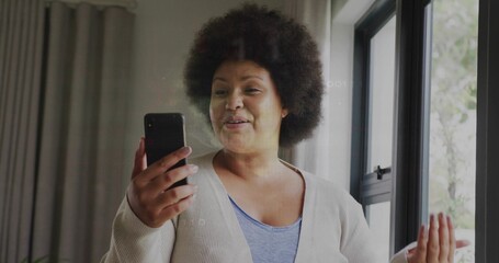 Holding smartphone, woman speaking, gesturing near window at home, wearing beige cardigan, blue top