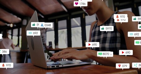 Typing female wearing sleeveless plaid top at cafe, using silver laptop with social bubbles