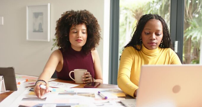 Diverse female coworkers pointing at printouts at home office table, annotating designs with marker