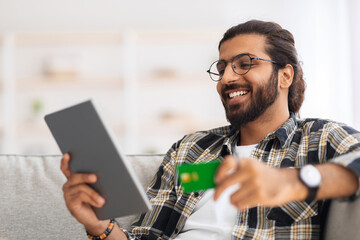 Closeup of handsome smiling arab guy banking online from home while lockdown, chilling on couch,...