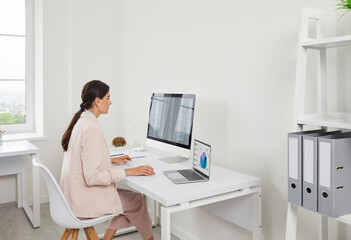Businesswoman analyst in office reviewing charts. She compares data on laptop and computer, preparing a report for work and audit in a bright setup. Clear data insights and work efficiency.