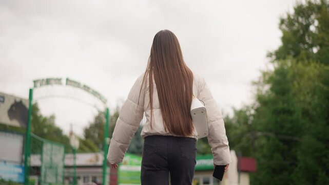 young woman walking toward park entrance under overcast sky, long braided hair in white jacket and jeans, walking away from camera past green gate and trees, shallow depth with soft bokeh, muted tones