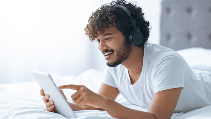 A young man lies on his stomach on a bed, wearing headphones and using a tablet. He smiles while interacting with the device. Sunlight enters the room through a window, creating a bright atmosphere.