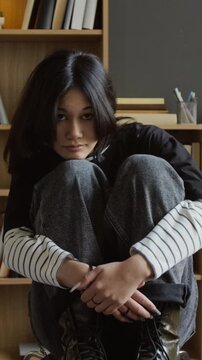 Vertical portrait shot of pensive teenage girl with long dark hair wearing emo styled outfit, sitting on chair in college therapists office while facing camera with calm expression