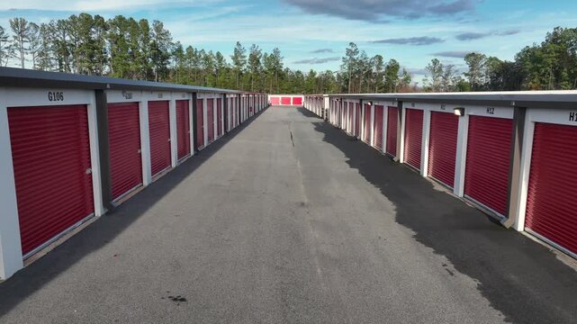 Aerial view of an American self-storage facility with long rows of red storage unit doors, clean asphalt lanes and soft daylight. Symmetrical, minimal industrial scene in U.S. Dolly backwards move