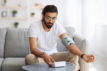 Long-haired, bearded millennial arab guy checking blood pressure at home, using tonometer while...