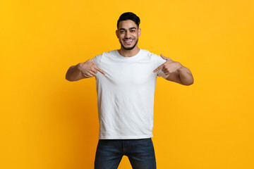 Cheerful arab guy pointing with both hands at his blank white t-shirt and smiling, posing over...