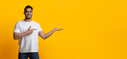 Handsome young arab guy in white t-shirt pointing at copy space for text or advertisement over yellow studio background and cheerfully smiling, panorama, showing nice deal or offer