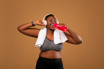 Sports Hydration. Portrait of curvy African American woman with white towel on neck drinking fresh water from bottle, wearing wireless headphones and sportswear isolated over brown studio background