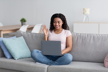 Fototapeta premium Online meeting, conference at home, be safe during social distancing and covid-19 outbreak. Smiling cute young african american female look at laptop and waving hand to webcam, living room interior
