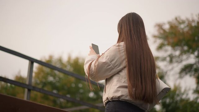 young woman with long hair standing on park bridge checking phone, scrolling messages, composing post, pausing to reflect against autumn trees and overcast sky candid lifestyle portrait with warm