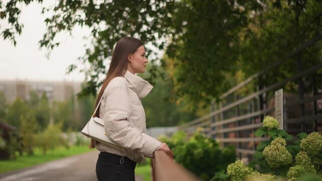 caucasian woman leaning on railing, autumn park path framed by greenery and hydrangea blooms, beige puffer jacket and white handbag, side profile portrait with cinematic shallow depth, overcast soft