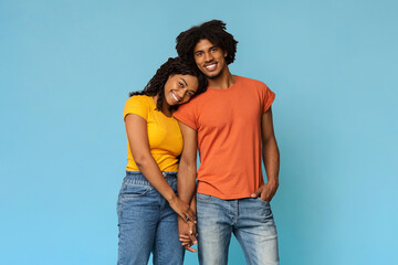 Loving young black lady bonding with her happy handsome boyfriend on blue studio background,...