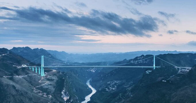 Aerial time-lapse of a majestic suspension bridge spanning a deep canyon with dramatic mountain scenery and soft dusk lighting, Huajiang Canyon Bridge in Guizhou Province, China