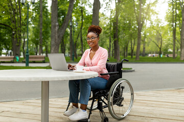 Cheerful handicapped black lady in wheelchair using laptop pc for online job at urban park, free...