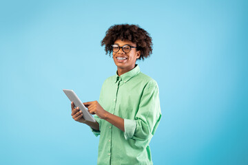 Online communiction. African american teen guy browsing web on tablet pc over blue studio background and smiling to camera. Funky student in glasses using touch pad, watching videos