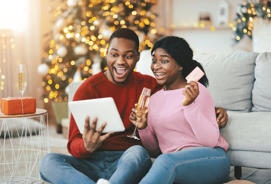A couple sits on the floor in their living room, looking excited as they use a tablet for online shopping. They hold a glass and a payment card. A decorated tree with lights is in the background. - Powered by Adobe
