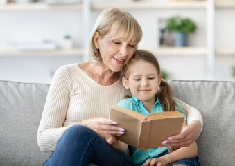 Naklejka premium Family Pastimes. Lifestyle portrait of loving caring granny reading interesting book together with her cute little granddaughter, hugging and sitting on the couch at home in living room