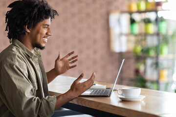 Smiling emotional curly young black man make video call on laptop, gesturing and communication at table in cafe interior. Online meeting, digital nomad, freelance, work remote and modern technology