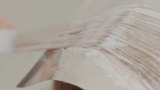 blonde white woman hair bleach application, stylist spreads lightening paste across foiled section with firm brush strokes, visible texture of product and saturated strand, closeup detail of chemical