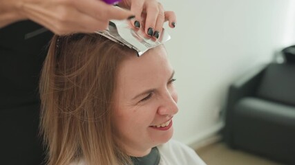 Caucasian woman smiling during hair highlighting session, colorist applying foil with comb, salon interior with black sofa, woman wearing protective cape, stylist hands with black nails placing foil,
