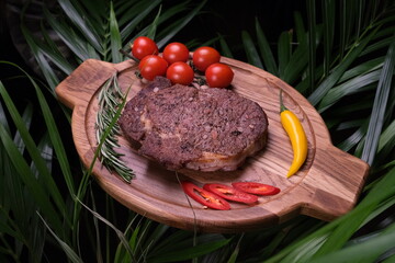 A juicy grilled beef steak served on an oval wooden board with cherry tomatoes, chili peppers, and rosemary against a tropical leaf background